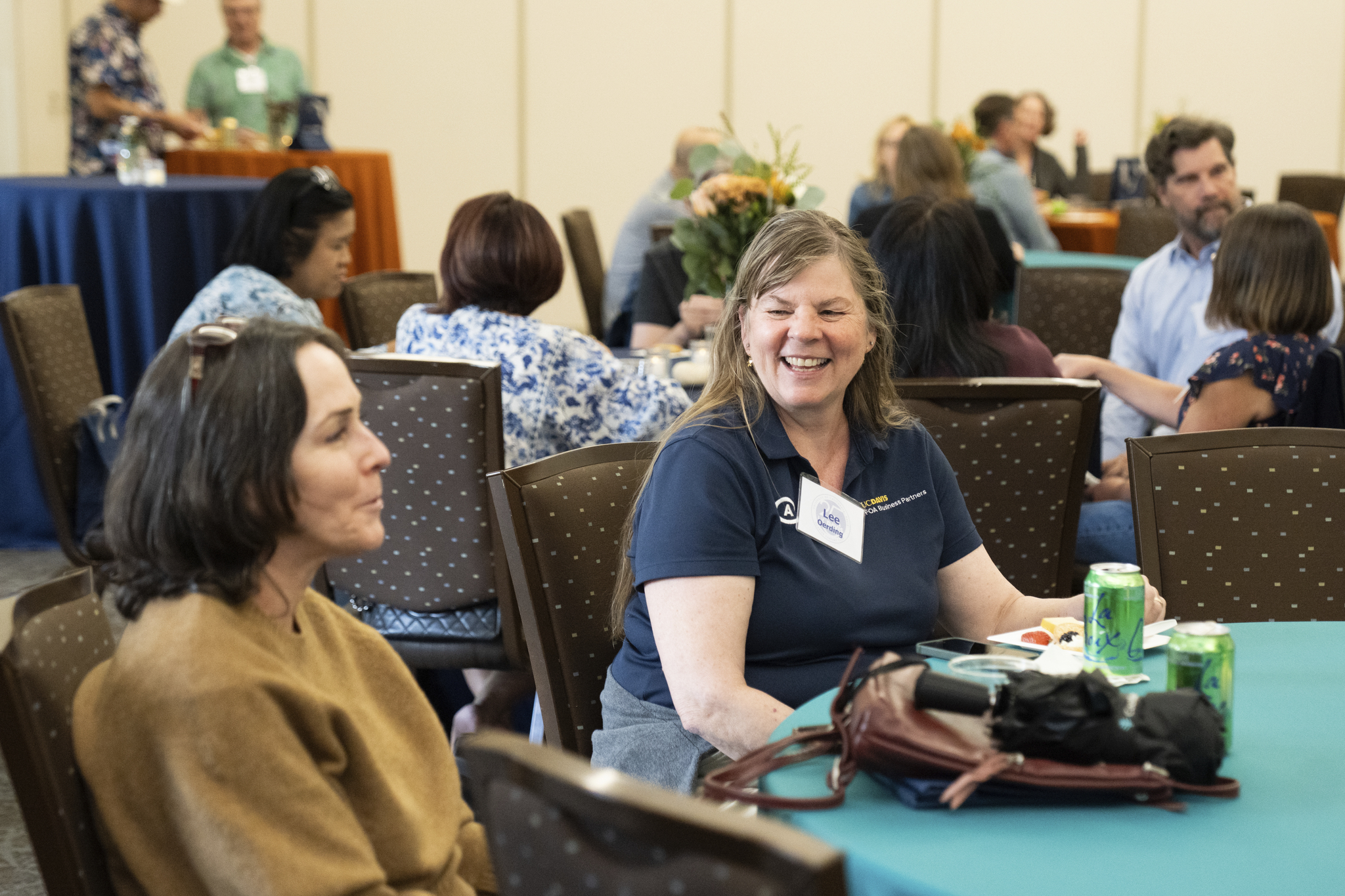Smiling woman with name tag at a conference table, attendees talking in background