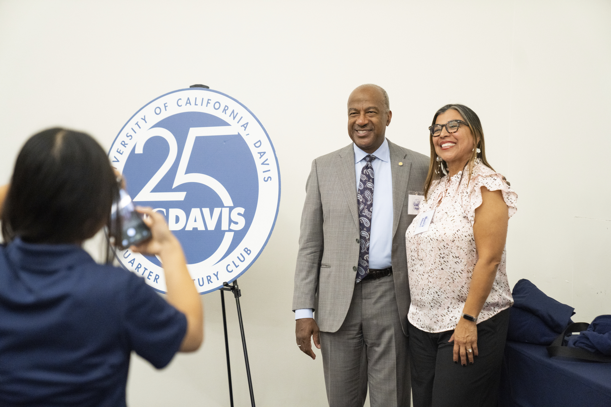 Two people smiling beside circular '25 Davis' anniversary logo at an indoor event