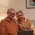 An older man and woman smile and sit together on a couch while looking at a laptop.